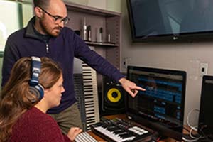 Student working at a DAW. Professor points to monitor. Music keyboards in view.