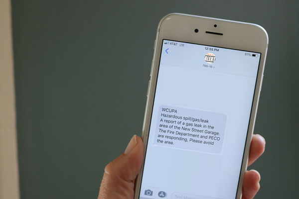 A photo of someone holding up a smartphone, with the screen showing a text from the WCU Alert system that reads: 'Hazardous spill/gas/leak. A report of a gas leak in the area of the New Street Garage. The Fire Department and PECO are responding. Please avoid the area.'